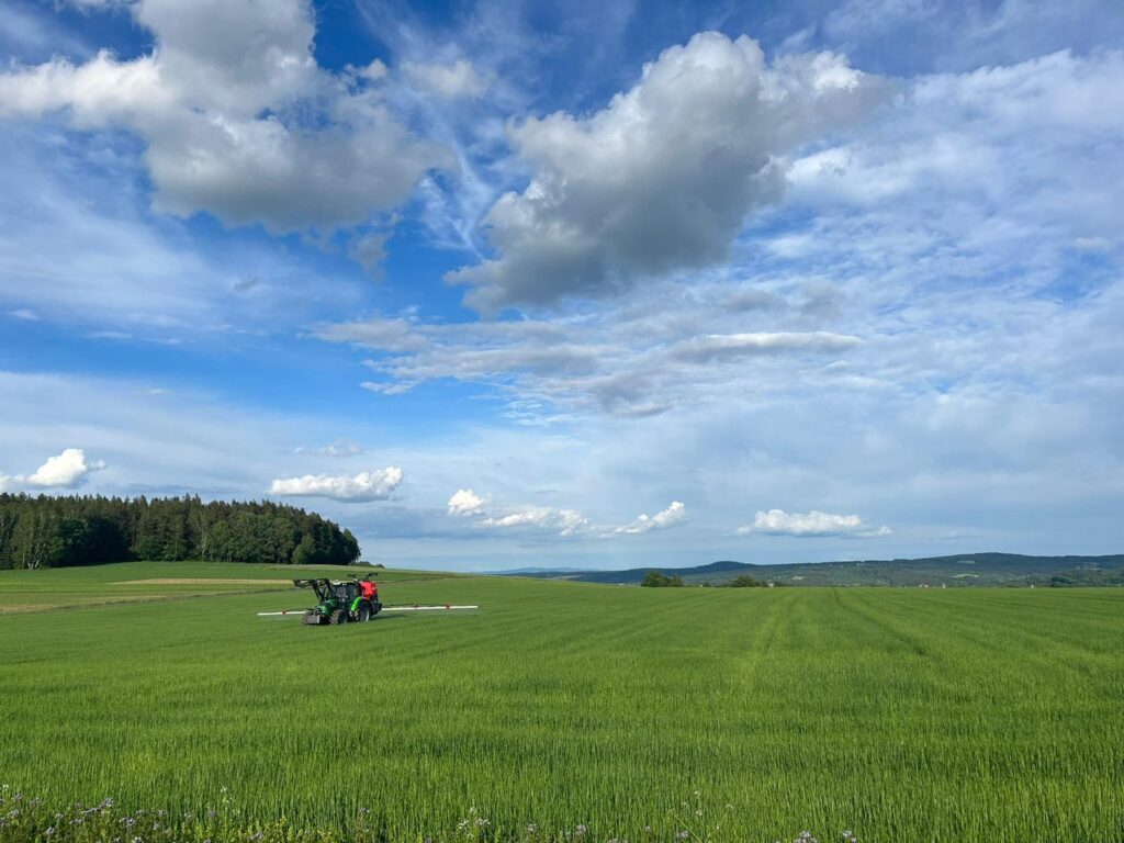 Ein Traktor bei der Ausbringung von Pflanzenschutzmitteln auf einem weiten Feld unter blauem Himmel.